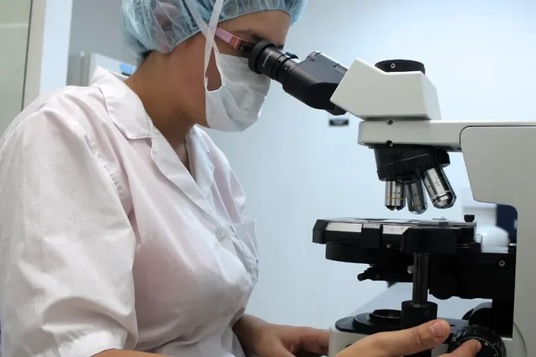Frau sitzt im Kittel mit Mund- und Haarschutz im Labor. Sie untersucht am Mikroskop die Verbindung zwischen Baum un Pilz